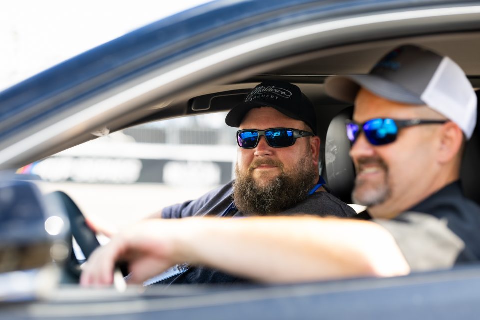 Image of an employee sitting in the passenger seat of a pace car. The driver has sunglasses on and is facing forward. 